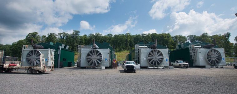 Four large box-like structures with huge fans sits across a gravel lot