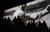 Silhouette of people from behind, seated, watching a black and while image of a worker projected on a wall