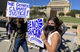 A young woman in a mask holds a sign that says end fracking.