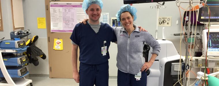 Pete Adams and Noe Woods stand arm in arm in an operating room wearing scrubs