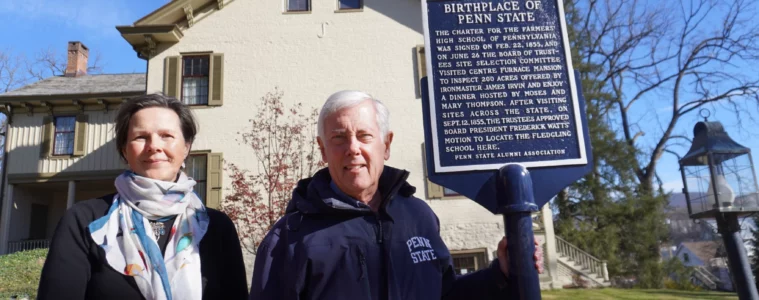 Mary Sorensen in a coat with a white scarf stands to the left of Roger Williams in a blue "Penn State Alumni" jacket next to a blue historical marker, with an historic house in the background