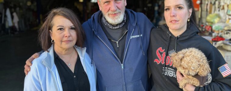 A man in a blue sweatshirt stands between two women. One is holding a small dog.