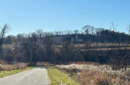 A facility can be seen on top of a hill in the distance, beyond a rural road and trees