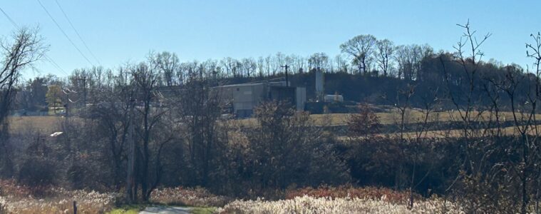 A facility can be seen on top of a hill in the distance, beyond a rural road and trees