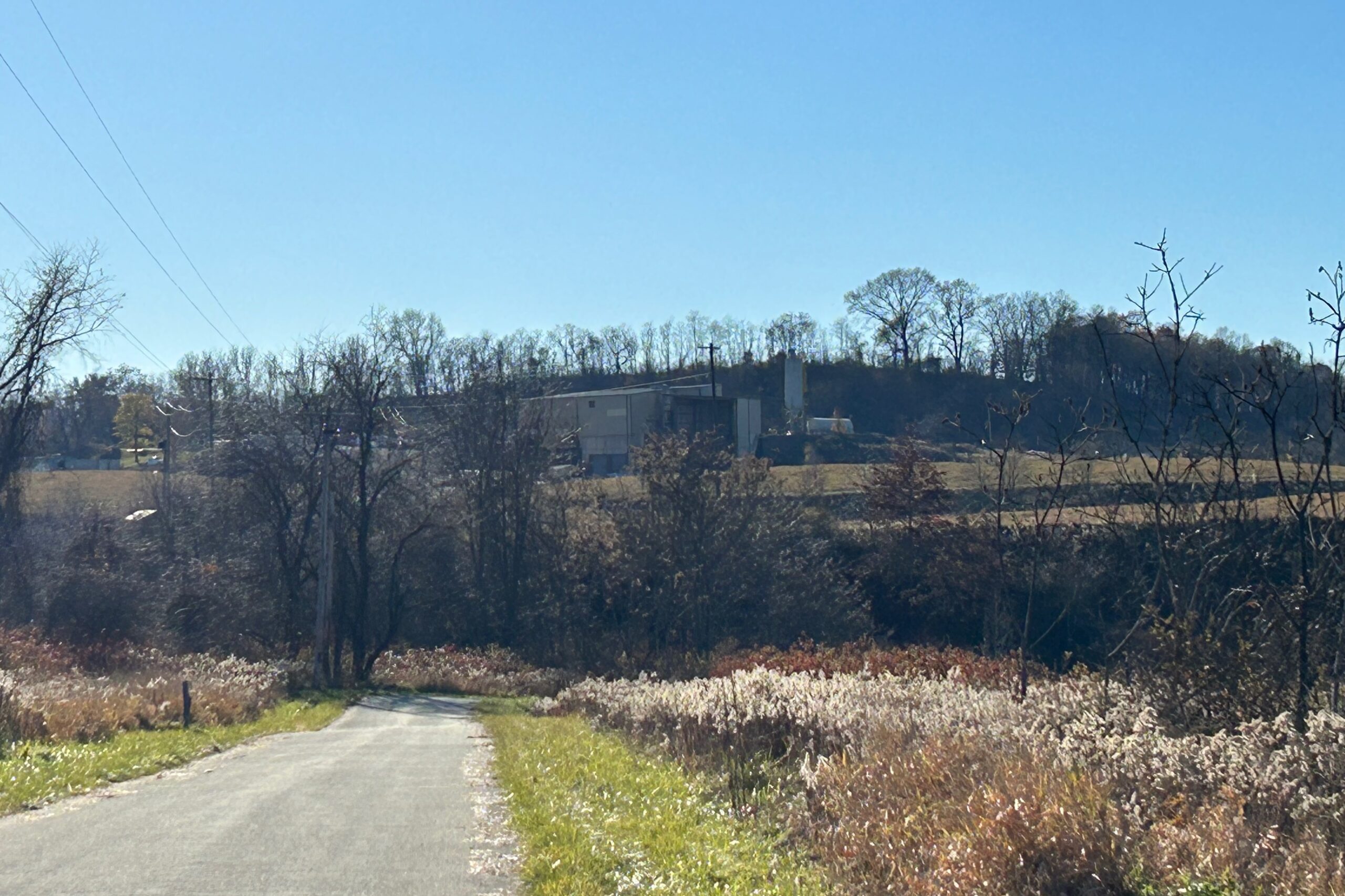 A facility can be seen on top of a hill in the distance, beyond a rural road and trees