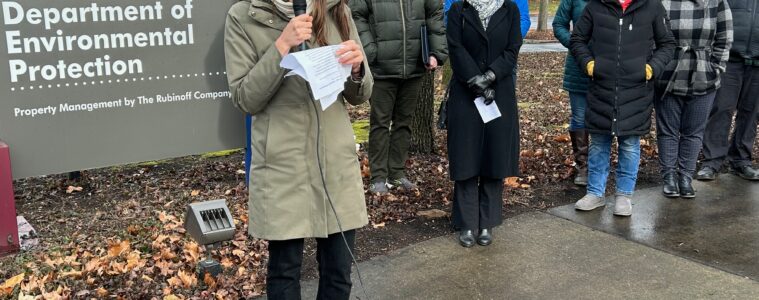 A woman stands with a microphone reading from a piece of paper she is holding, in front of a sign that reads "Department of Environmental Protection" while others in the demonstration look on behind her.