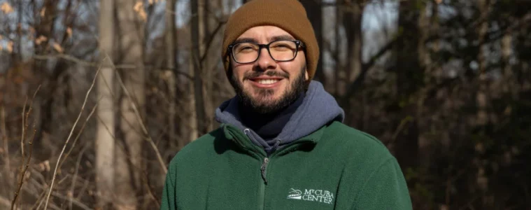 A man with a brown knit hat and green hoodie with glasses and a beard stands in a forest