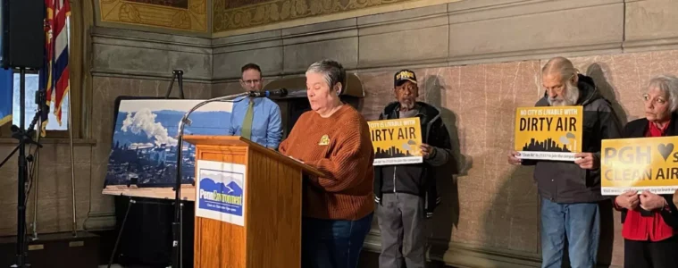 A person stands at a podium, while 3 others line up behind her holding signs that read "Dirty Air."