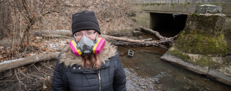 A woman stands in a stream in front of a culvert going under a home. She is wearing a winter coat and hat with a respirator.