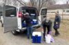 Three people unload blue coolers from a van