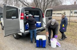 Three people unload blue coolers from a van