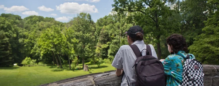 Two people stand on a stone bridge looking at a park with green space and trees