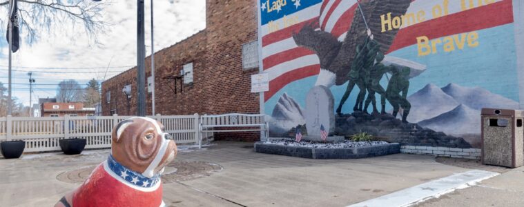 A statue of a bulldog painted red, white and blue sits along the sidewalk with a patriotic mural in the background