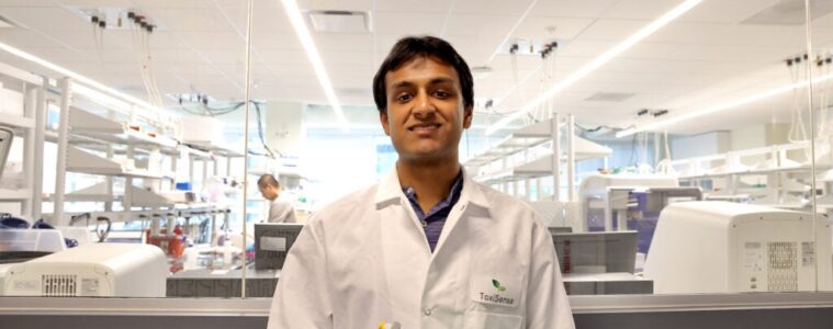 Aravind Krishnan is wearing a white lab coat standing in a la