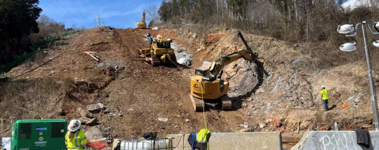 Construction equipment on a cleared hillside, with a worker in the foreground, behind concrete barriers