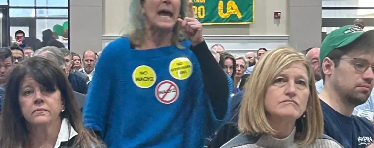 A woman in a blue shirt with protest pins stands in an audience yelling. In front of her are two ther women wearing pins.