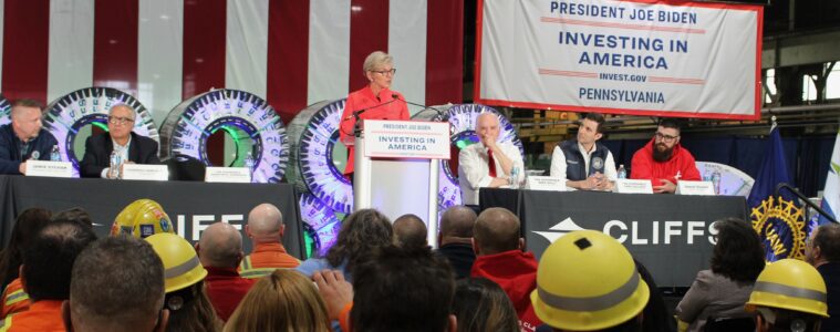 A woman stands at a podium with a row of men sitting at a table behind her, and a seated crowd in front, including workers in orange jackets and yellow hardhats.