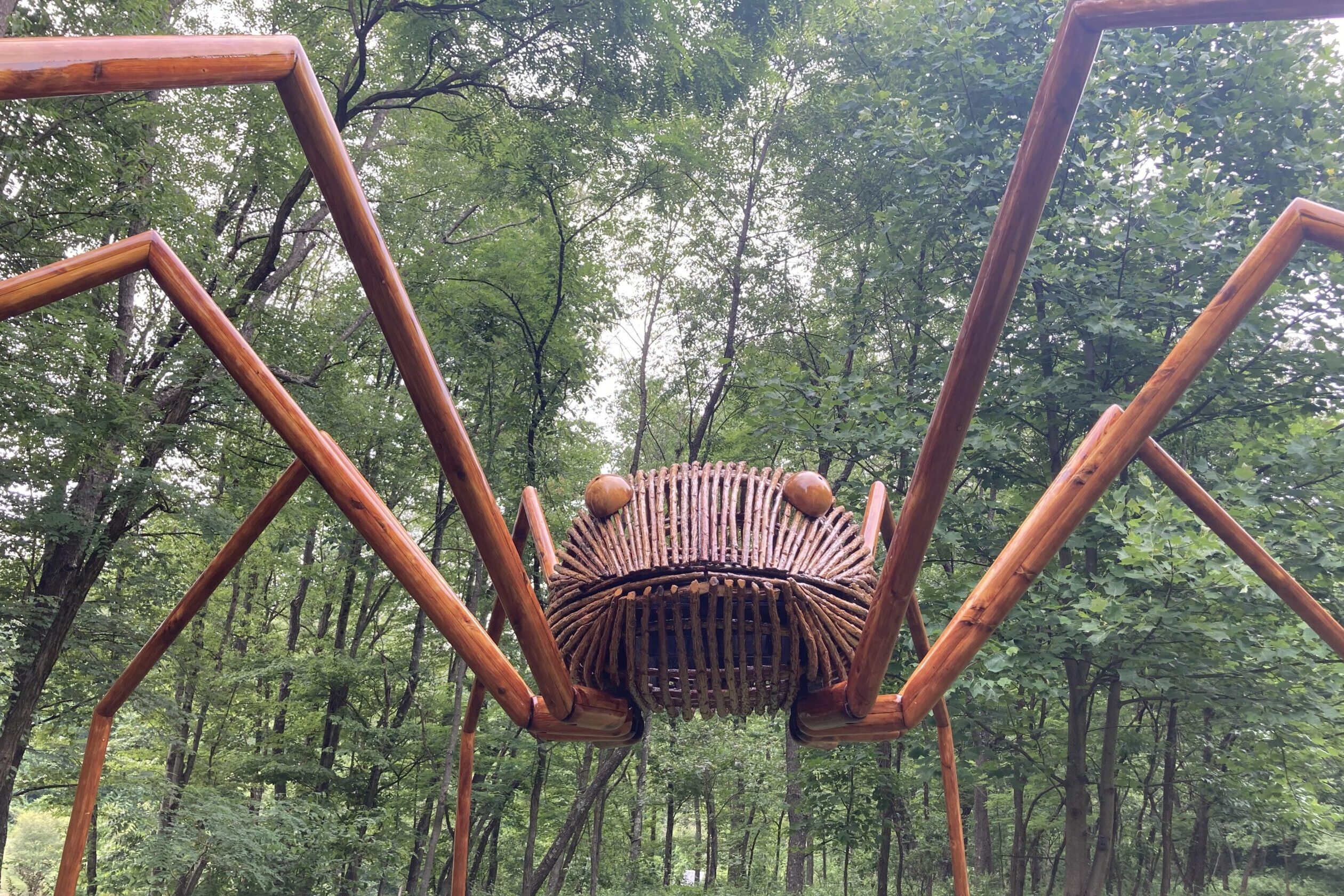 a giant daddy long legs sculpture made of wood set against green trees
