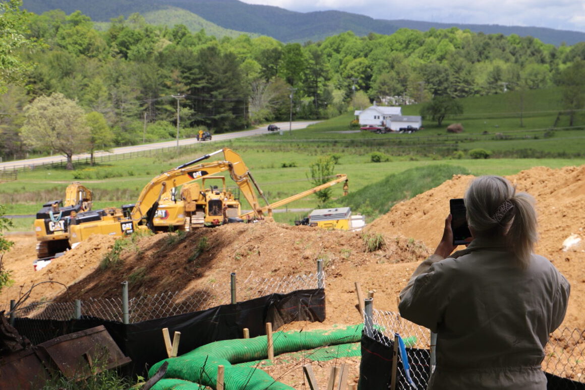 A blond woman with a pont tail photographs a yellow construction vehicle moving dirt along the Mountain Valley Pipeline