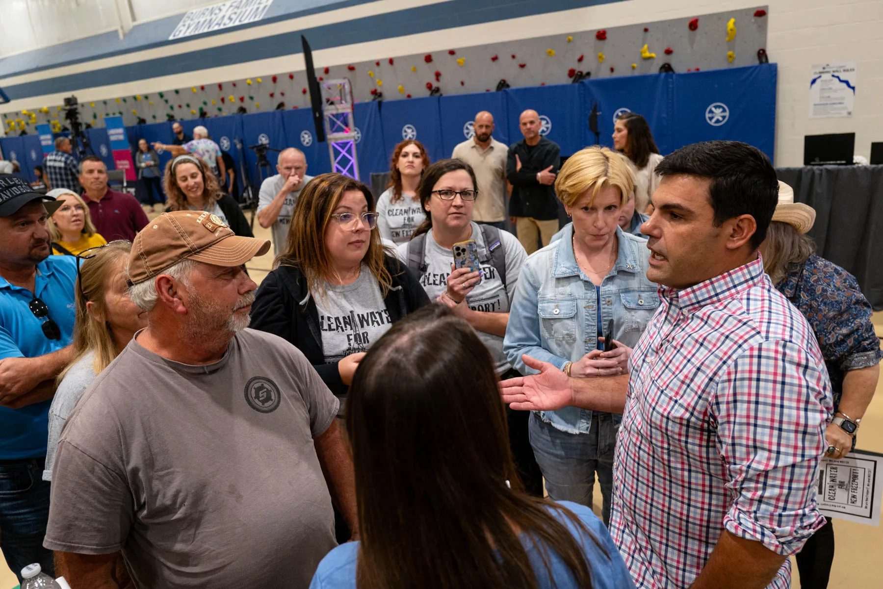 People stand around a man in a plaid shirt at a public meeting