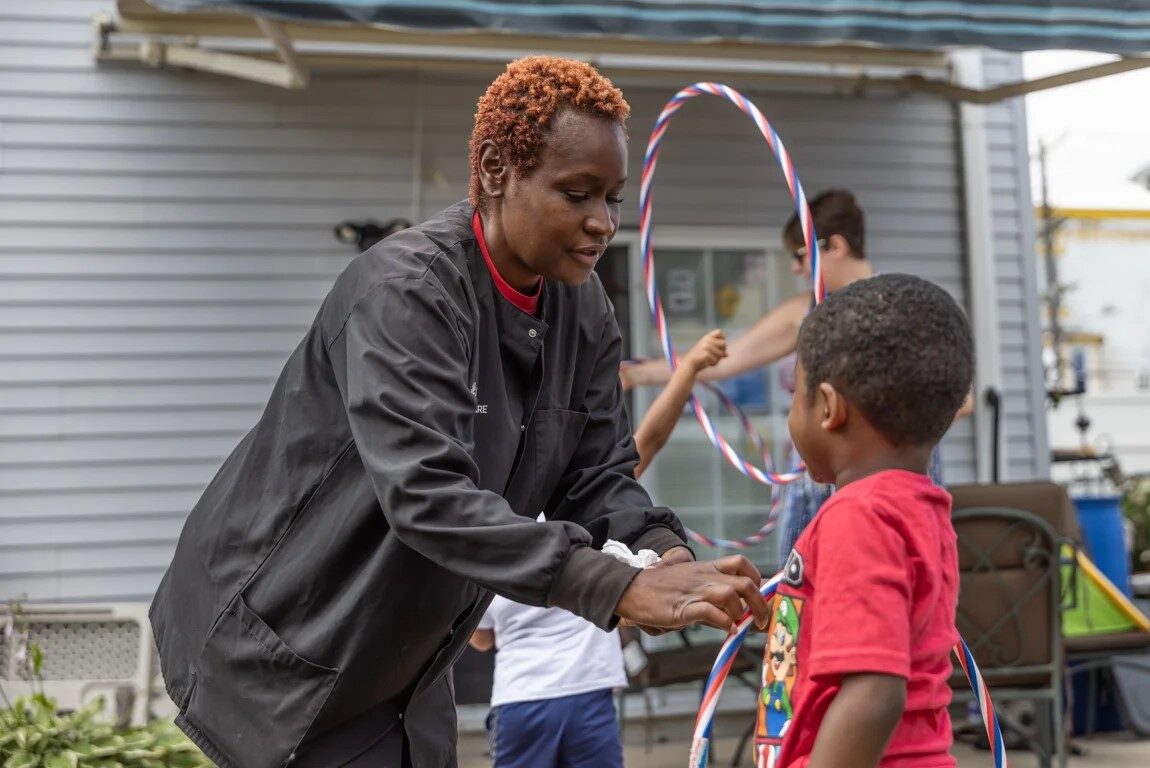A woman with short, curly red hair stoops over to hand a small child a hula hoop.