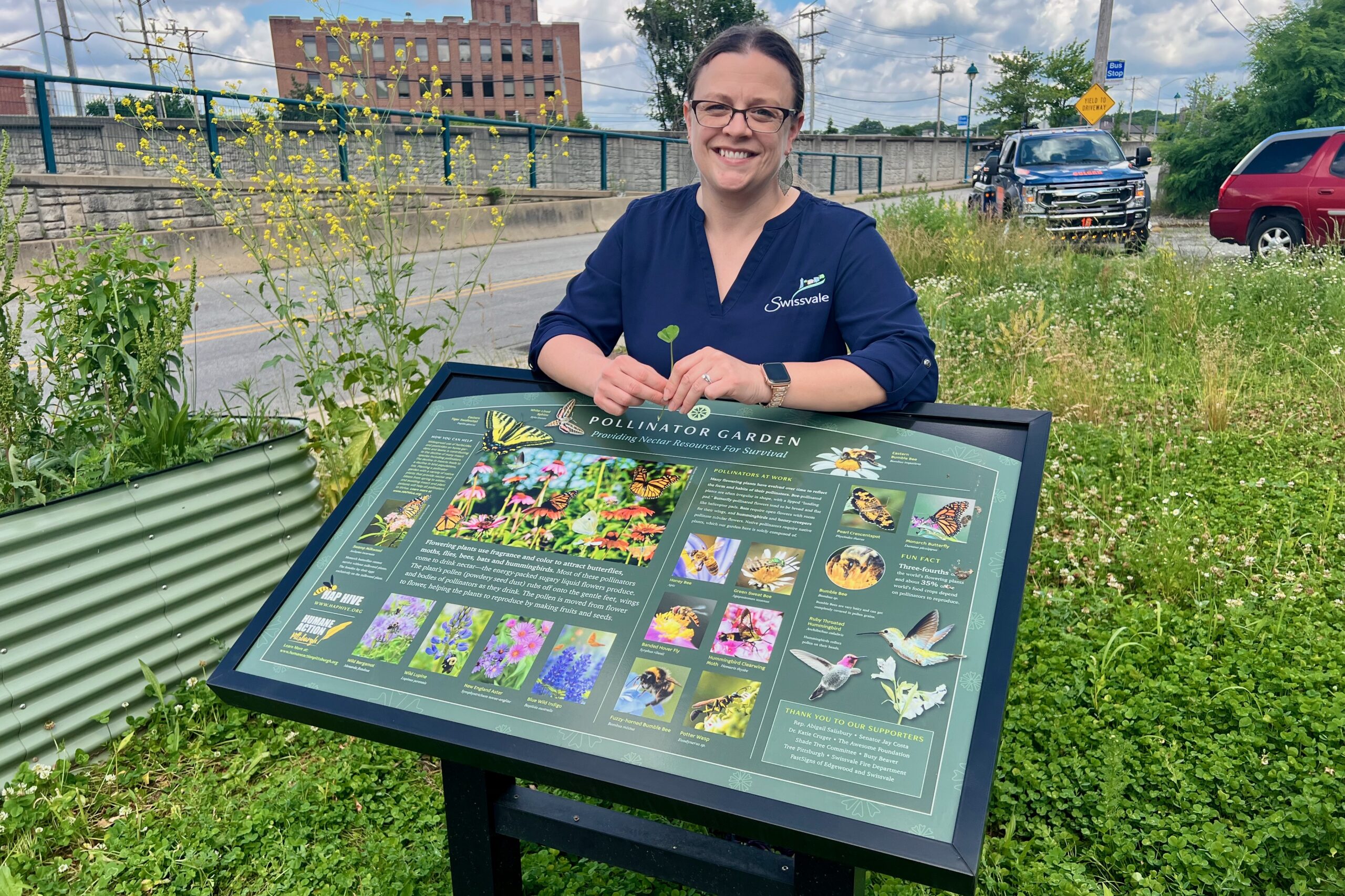 A woman smiles and rests her arms on a sign depicting flowers and insects at the edge of an outdoor garden
