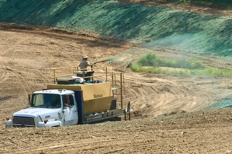 A large truck with a person in the bag spraying seeds soon a dirt field.