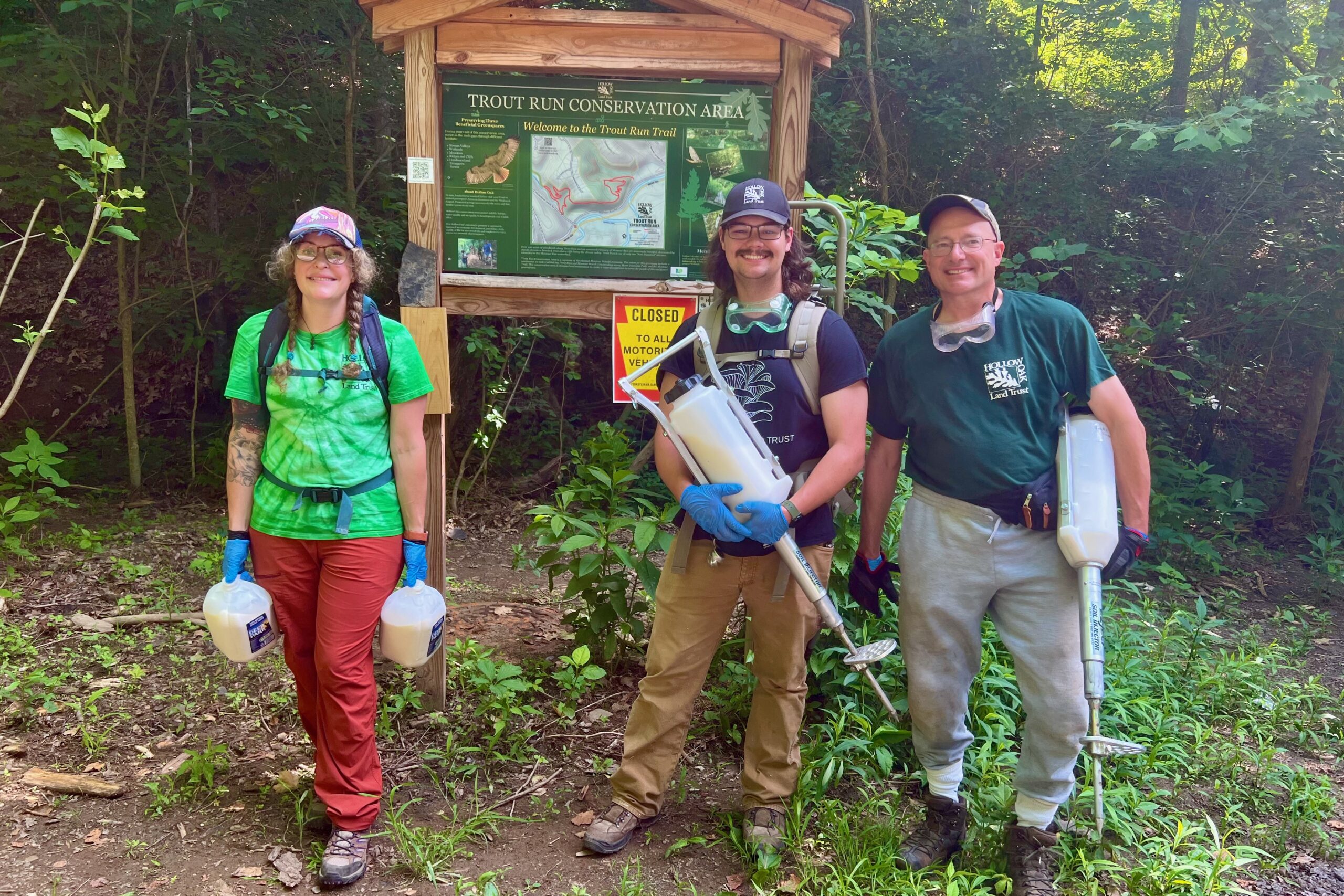 Three Hollow Oak Land Trust workers stand in front of a sign that says, "Trout Run Conservation Area." They are wearing backpacks and equipment, while carrying jugs and soil injectors of treatment. They are surrounded by wooded scenery.
