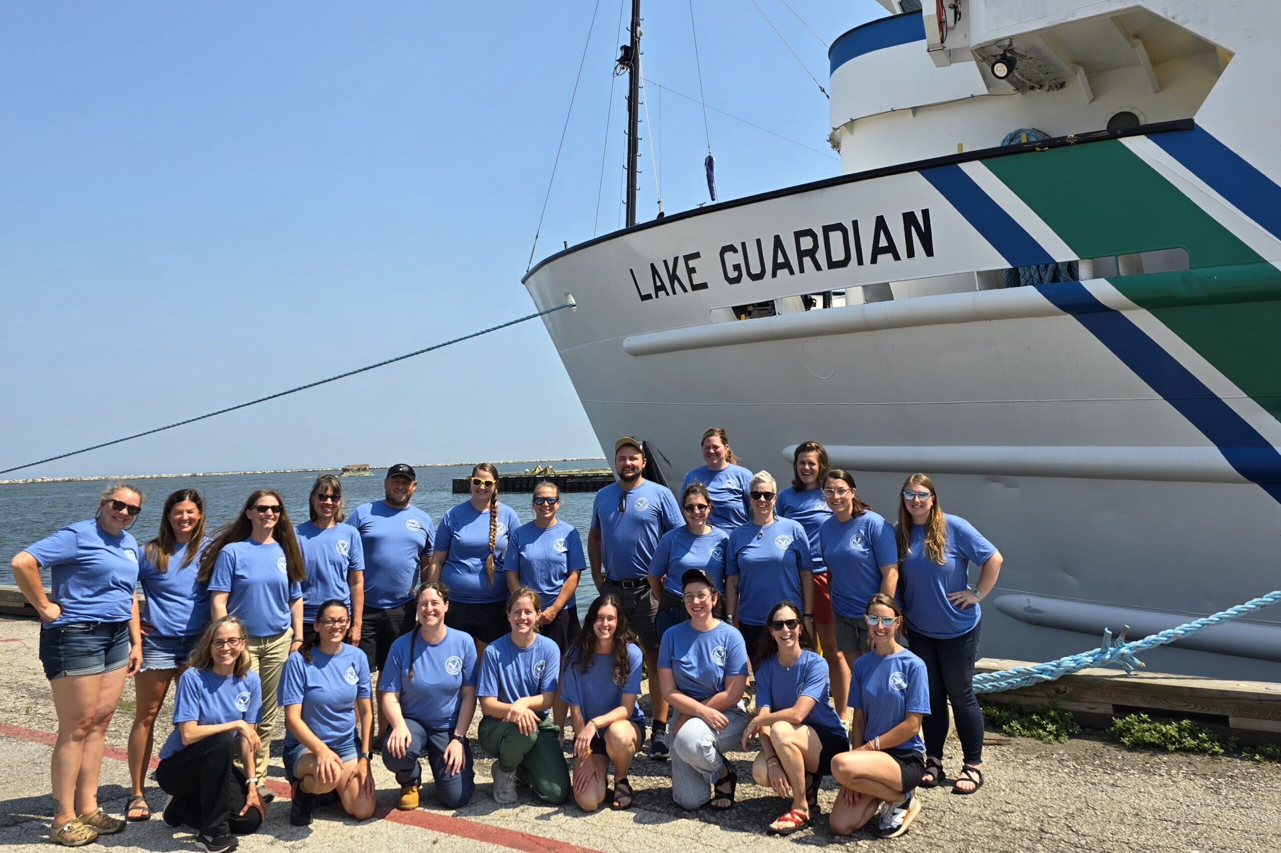 A group of 15 people in blue shirts pose in front of a big ship.