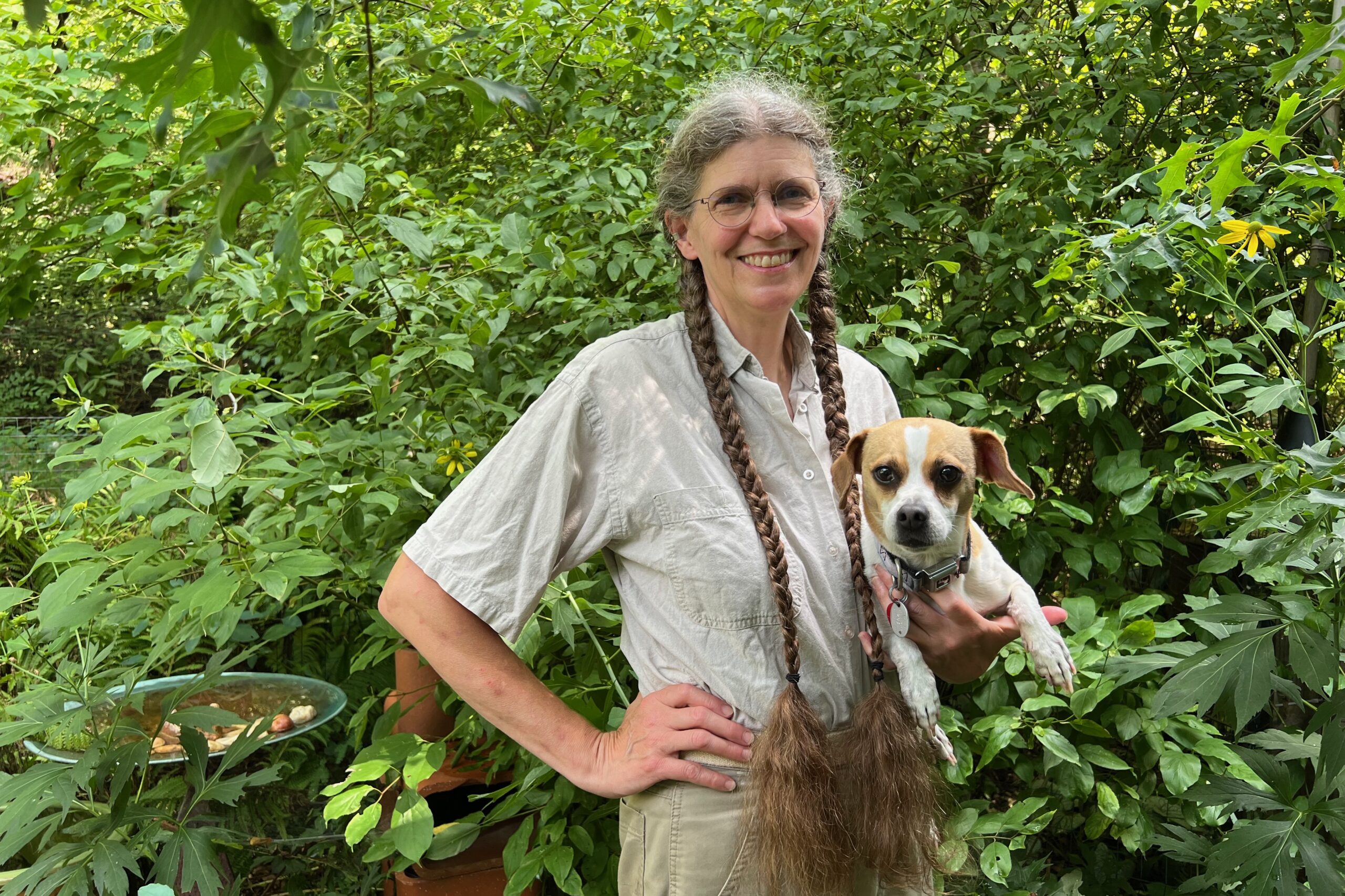 A woman stands with long braids stands holding a dog, with greenery in the background.