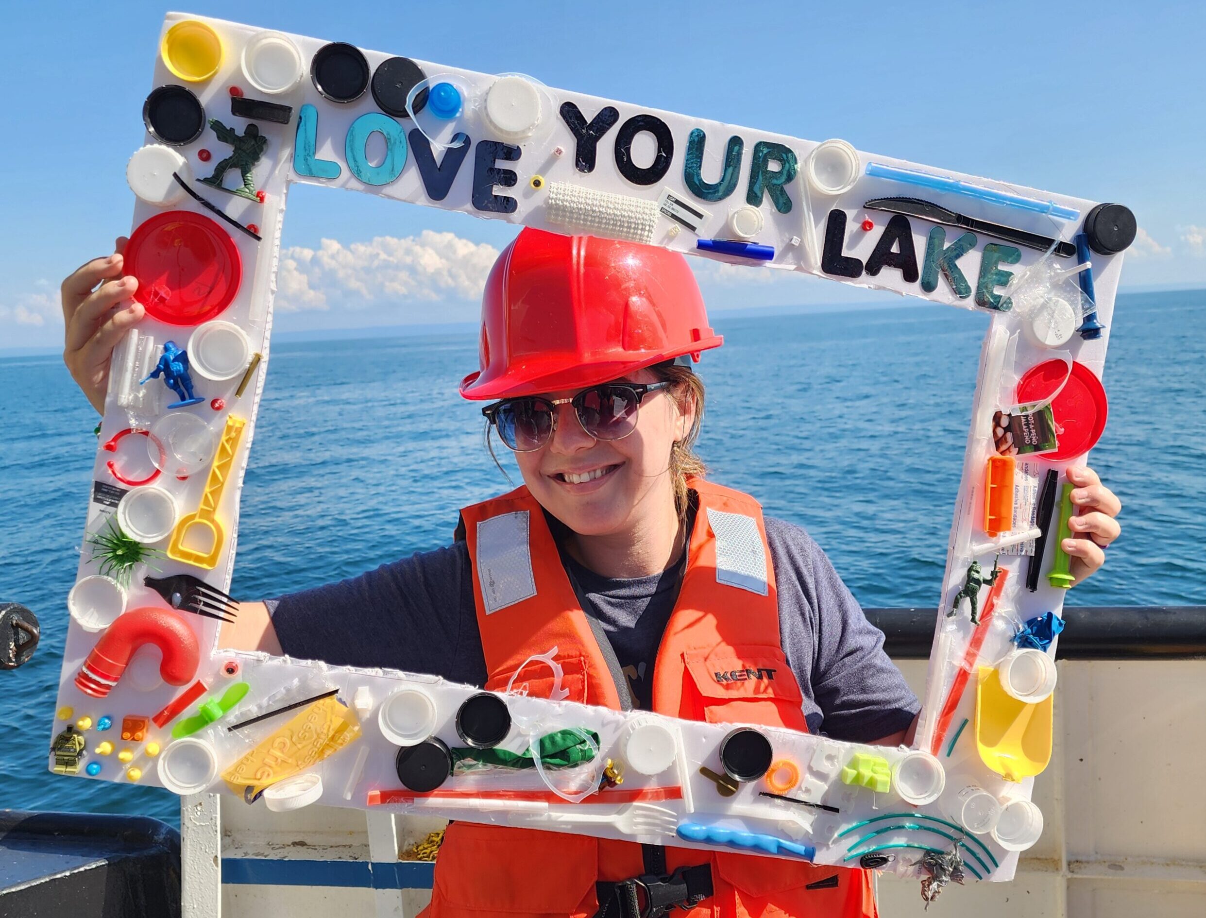 A woman in a red hard hat and orange vest smiles through a large frame decorated with multi-colored pieces of plastic.