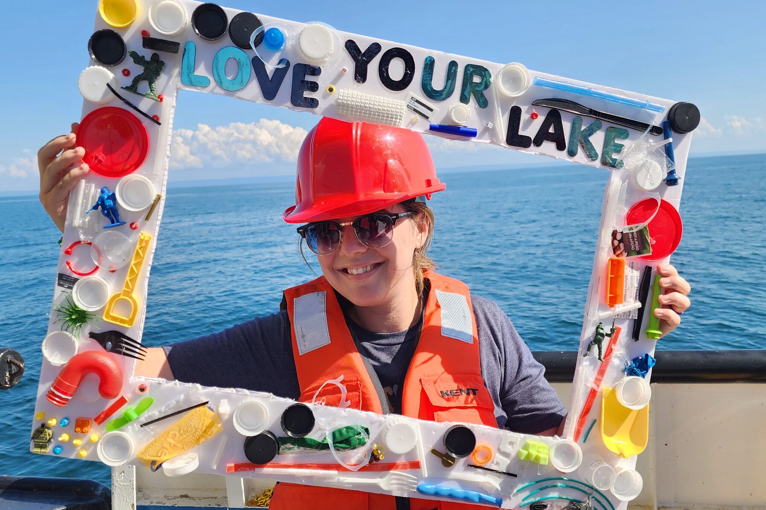 A woman in a red hard hat and orange vest smiles through a large frame decorated with multi-colored pieces of plastic.