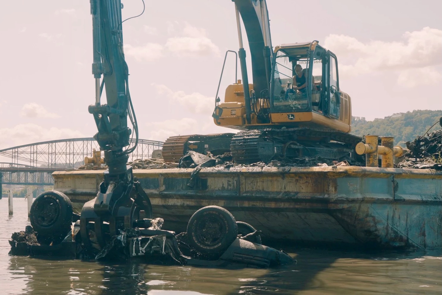 A large excavator on a barge pulls a car from the river.