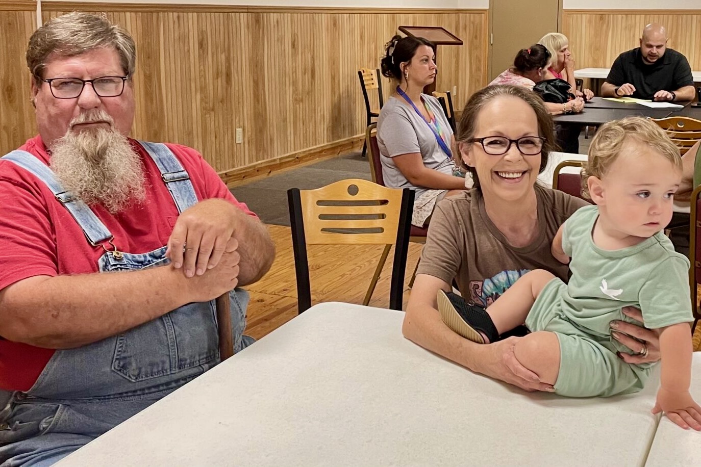 A man with a long beard and overalls sits at a table with a woman holding a toddler who sits on top of the table.