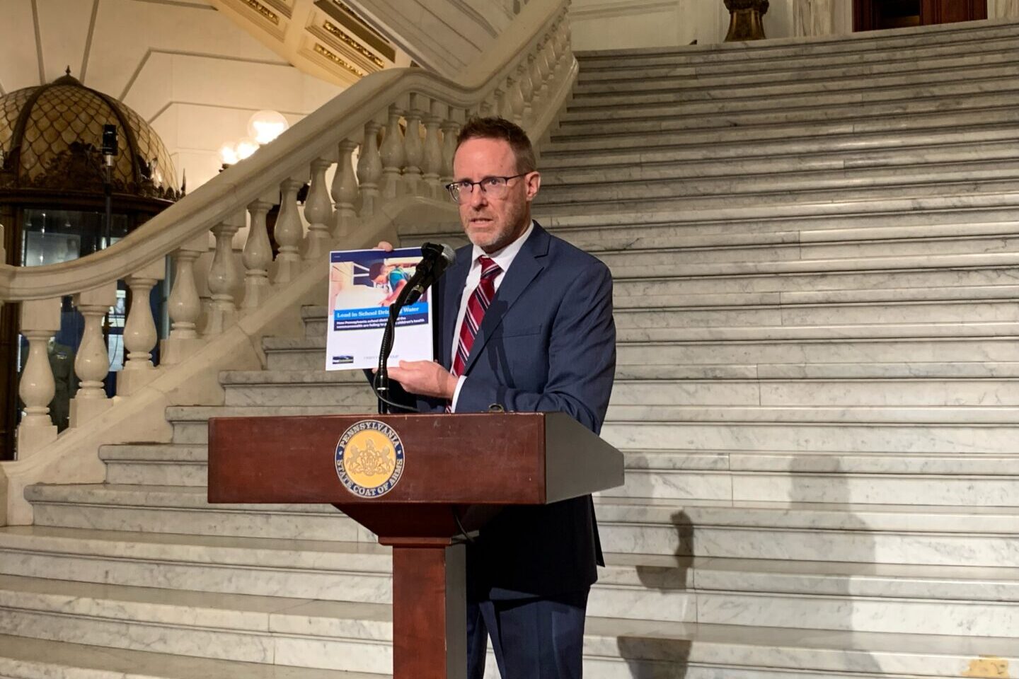 A man stands at a podium at the bottom of steps inside the state capitol holding a report.