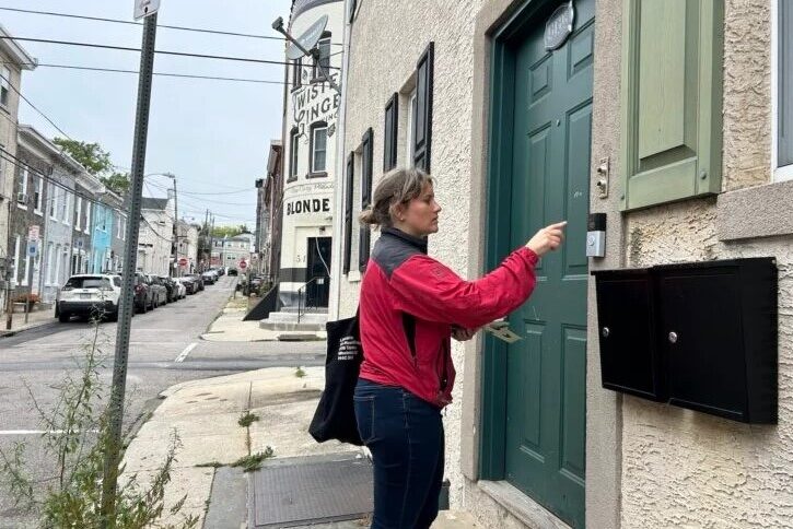 A woman with blond hair in a red jacket knocks on the door of a house