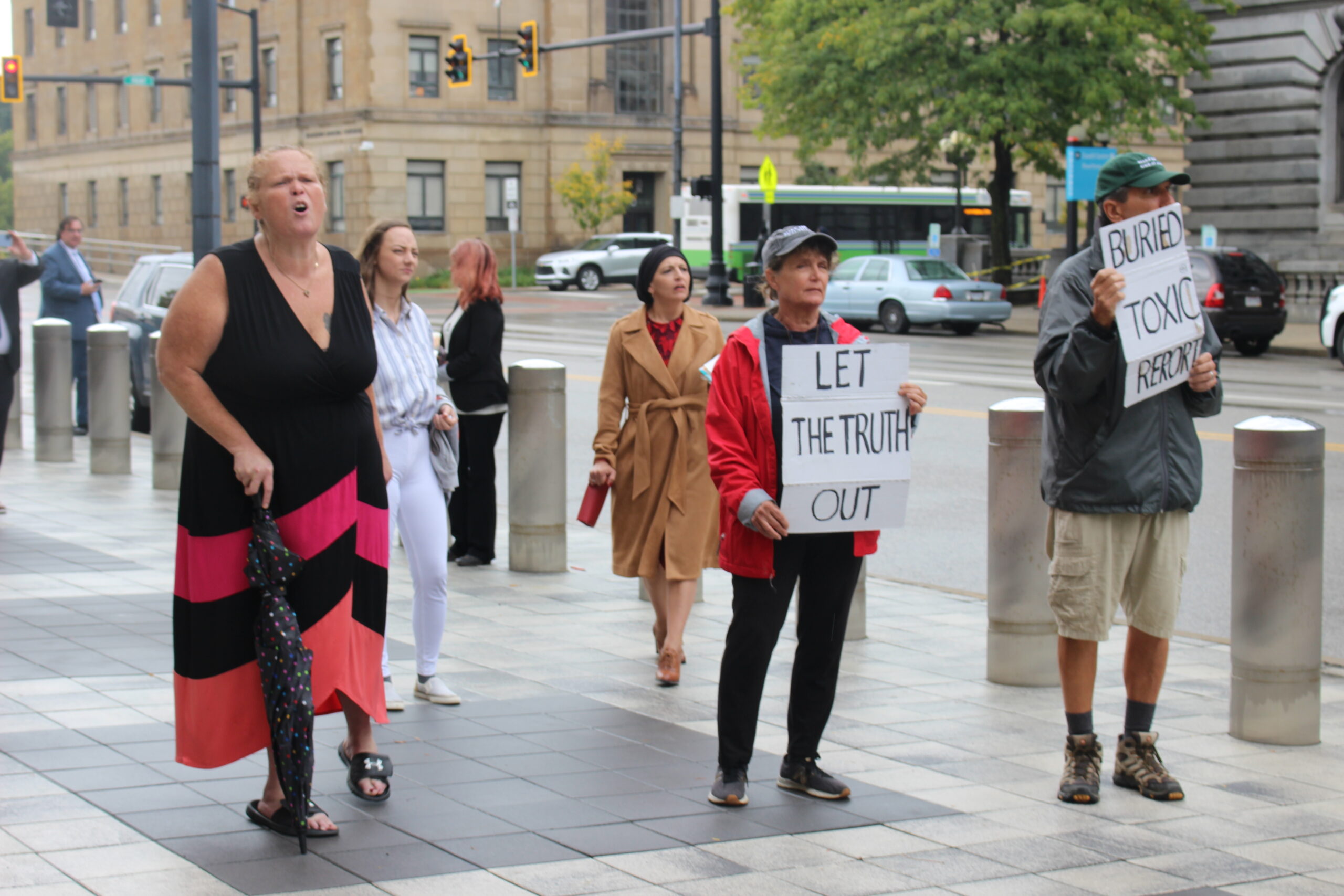 People standing on the sidewalk, one holding a sign that says "Let the Truth Out."