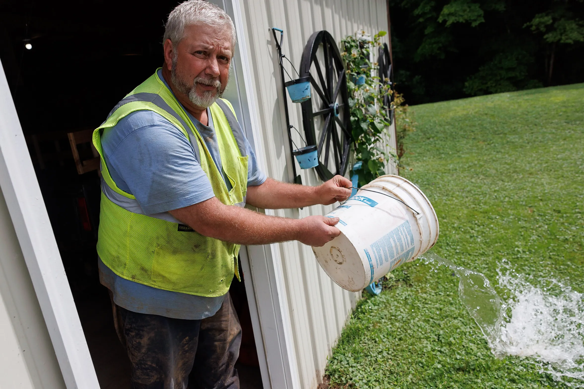 A man, standing in a doorway wearing a yellow safety vest, throws water from a white bucket.