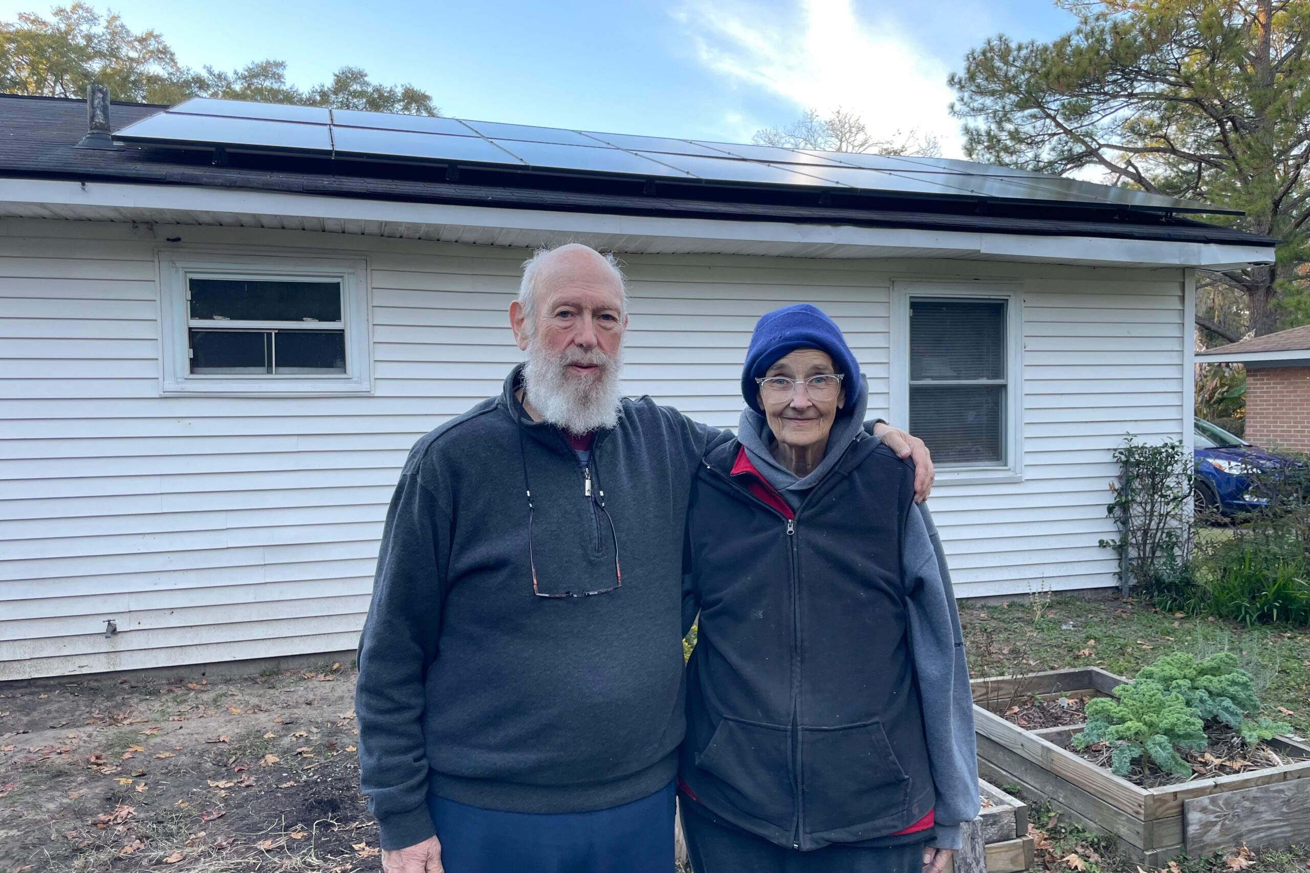 An elderly man and woman stand in front of a modest, one-story house with solar panels on the roof.