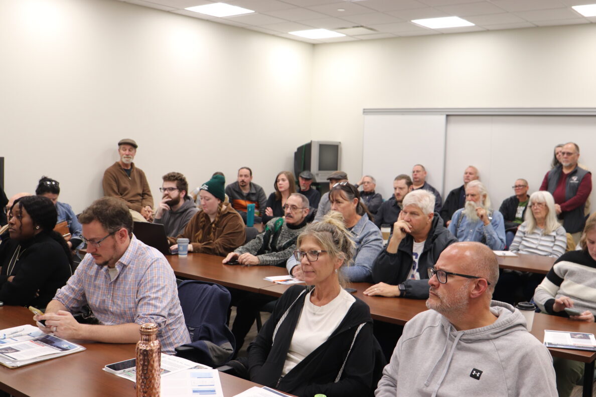 People sit in rows behind desks to listen to a speaker at the front of the room.