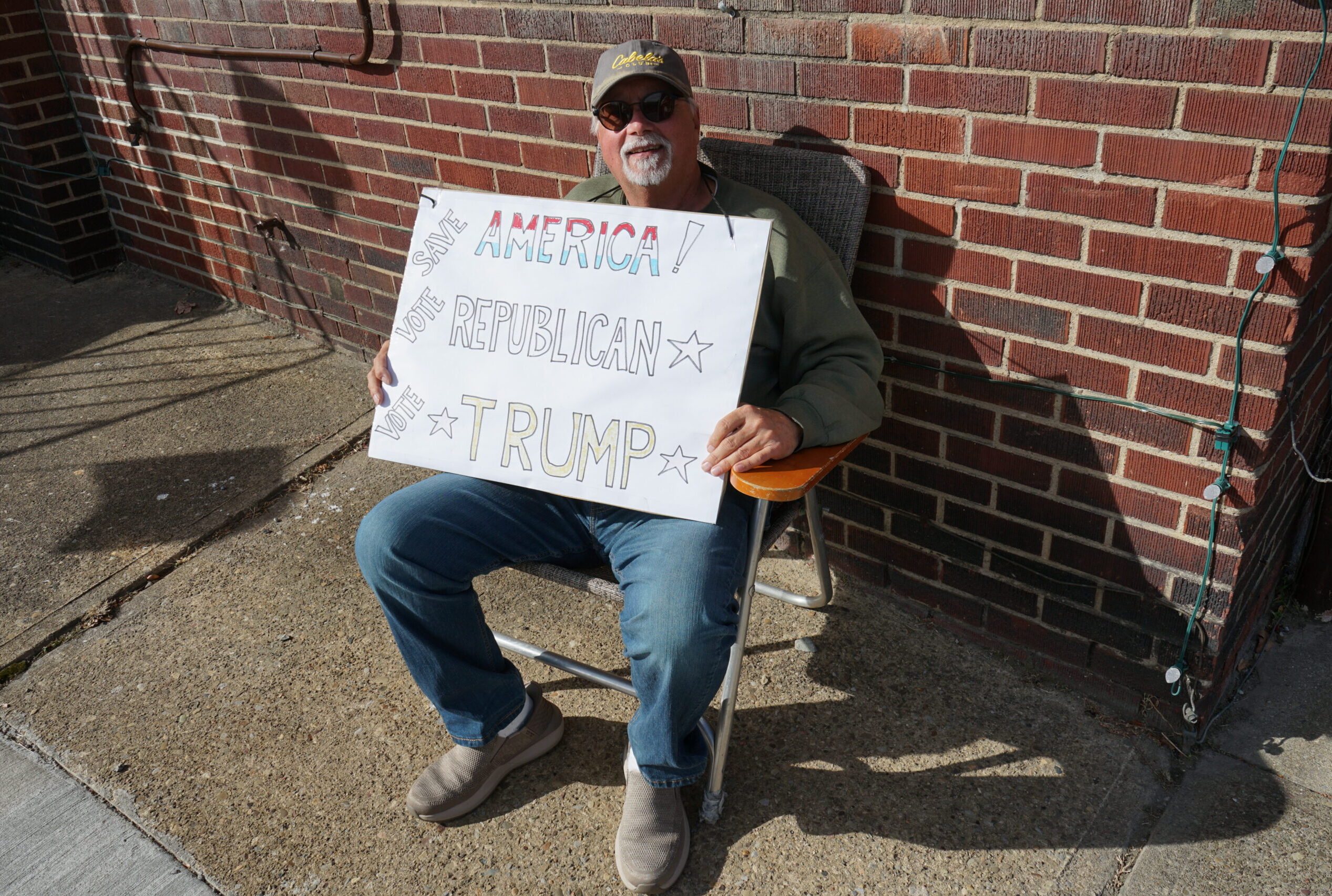 A man sits in a chair wearing a baseball hat and sunglasses, holding a sign in support of Trump