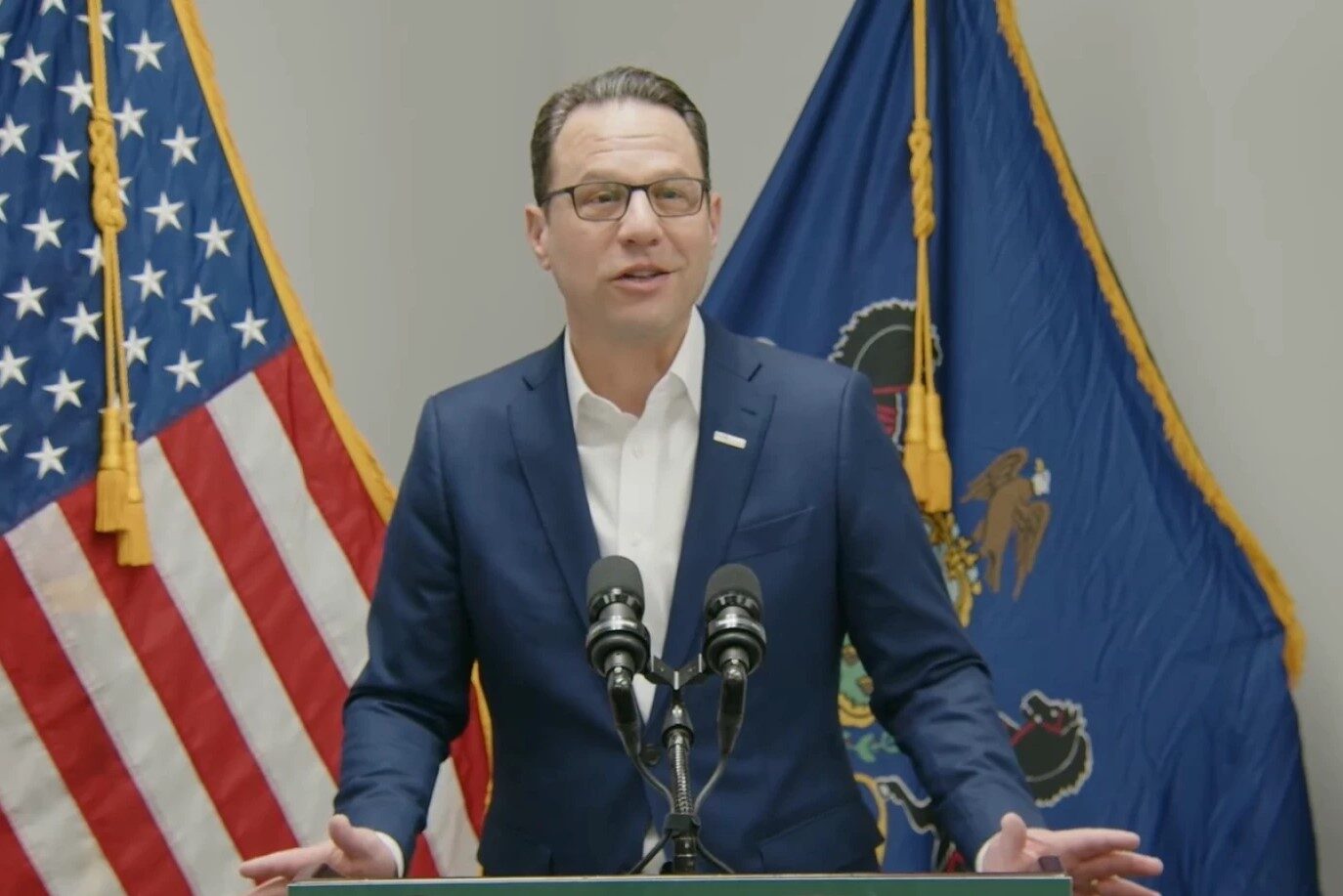 Governor Josh Shapiro stands behind a podium with the Pennsylvania and American flags behind them.