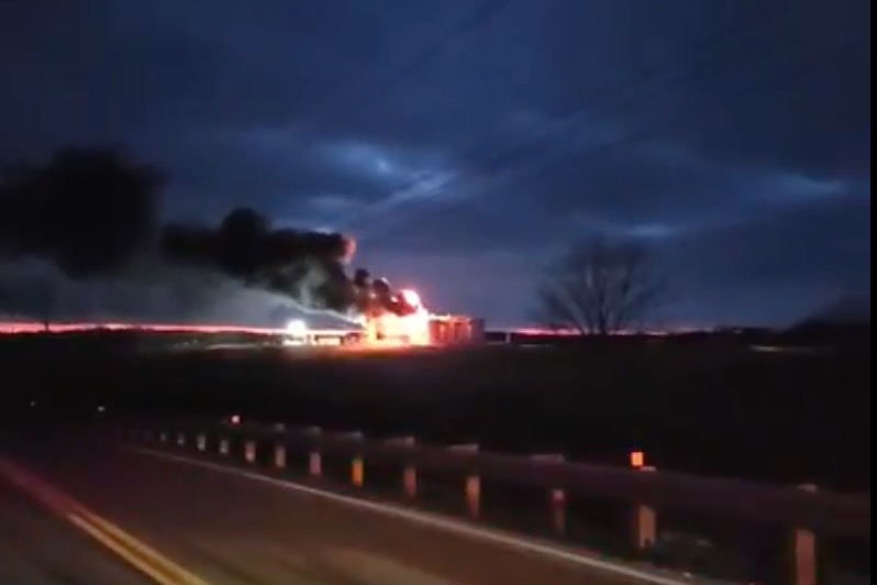 A structure on fire across a road with thick black smoke under a dark cloudy sky.