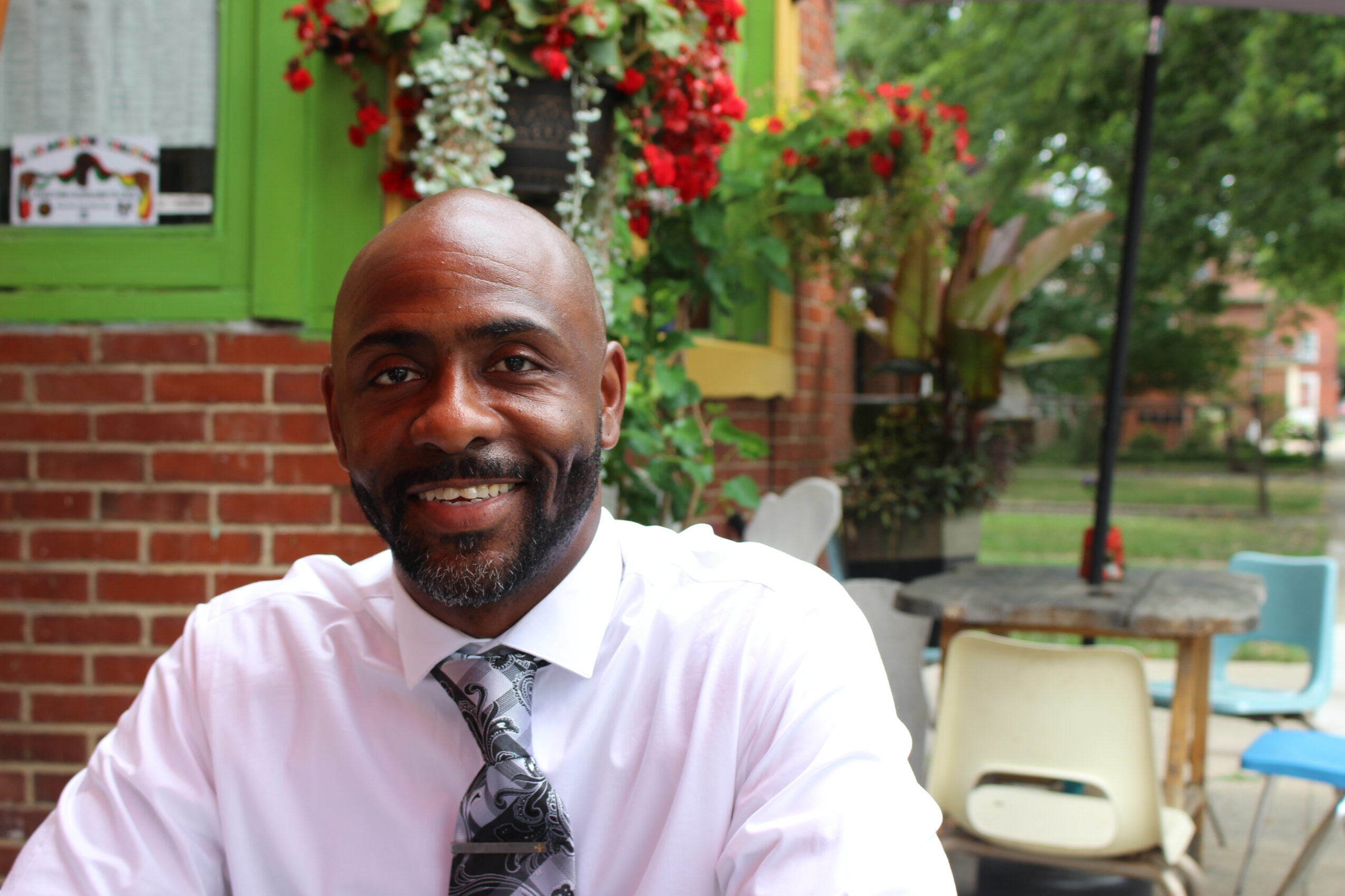 A man up close, sitting at a cafe with red flowers behind him.