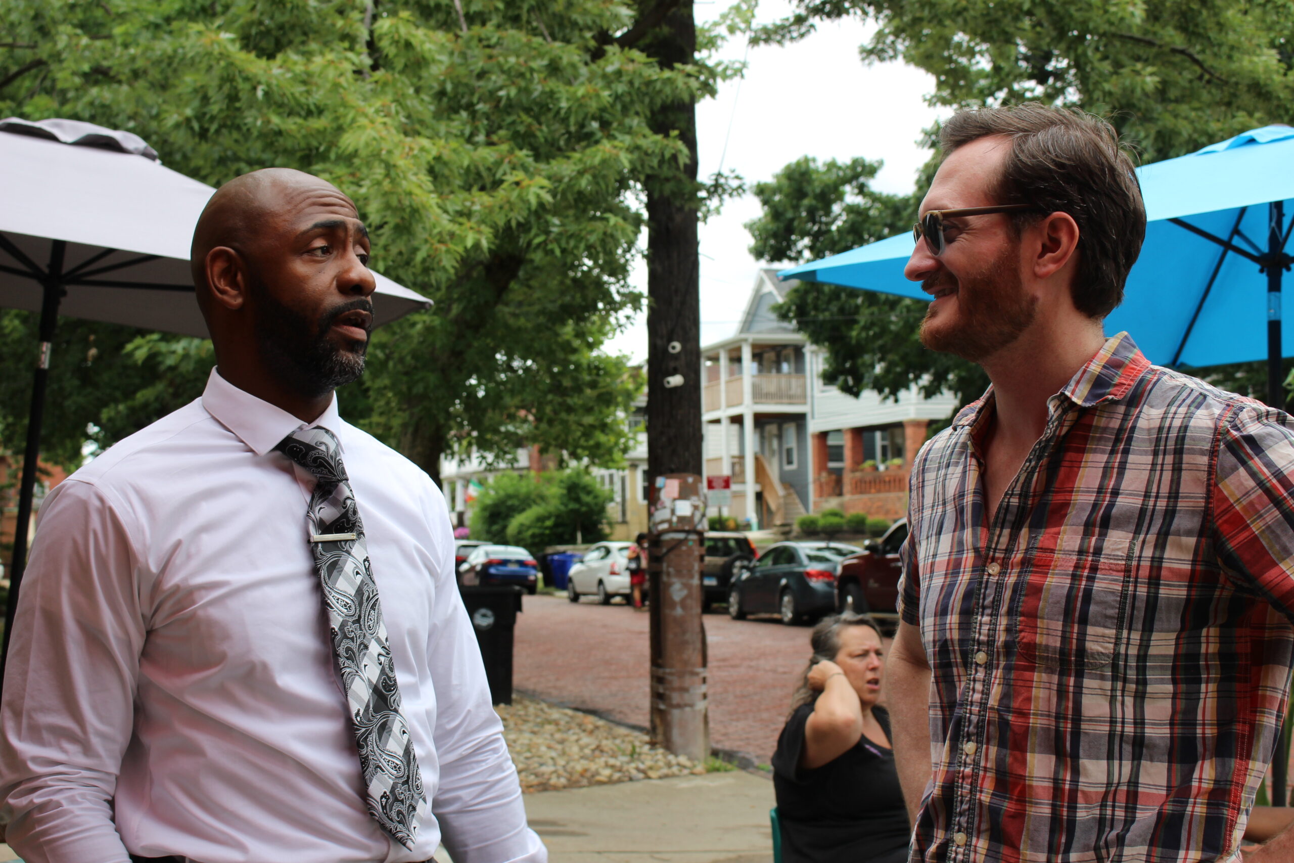 Two men standing, talking outside a cafe, colorful umbrellas can be seen