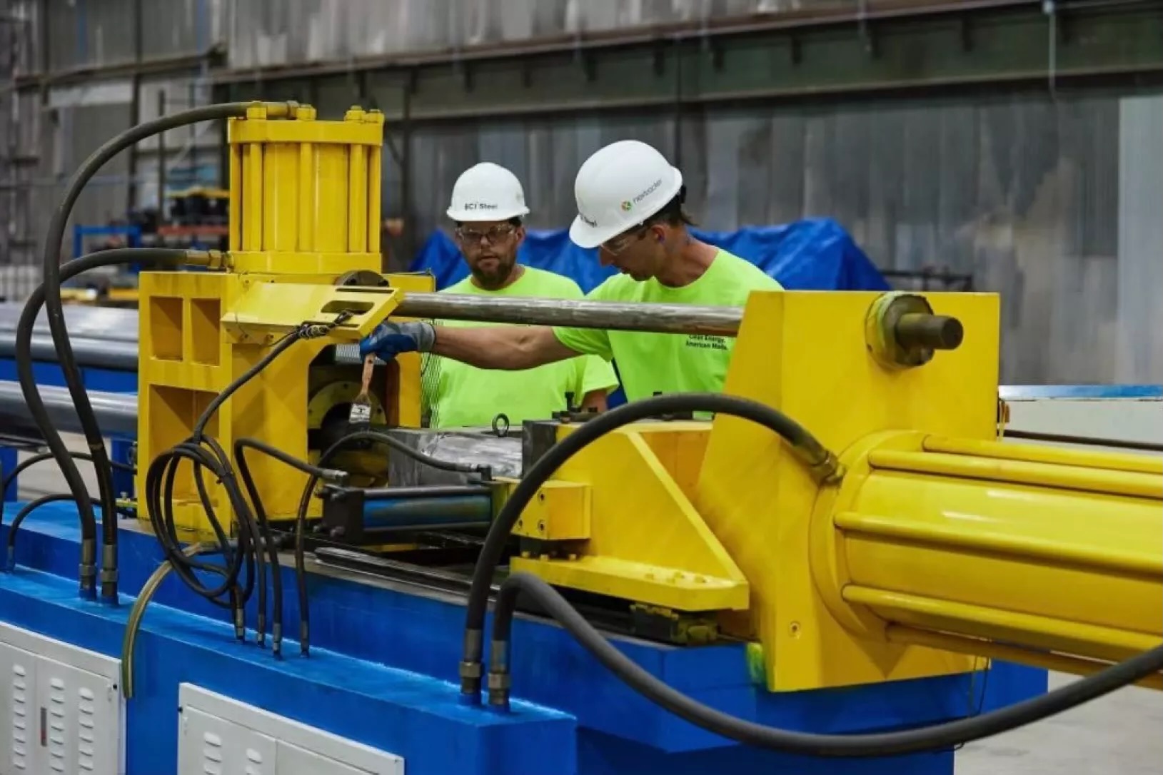 Two workers in white hard hats and bright green shirts work on a piece of equipment