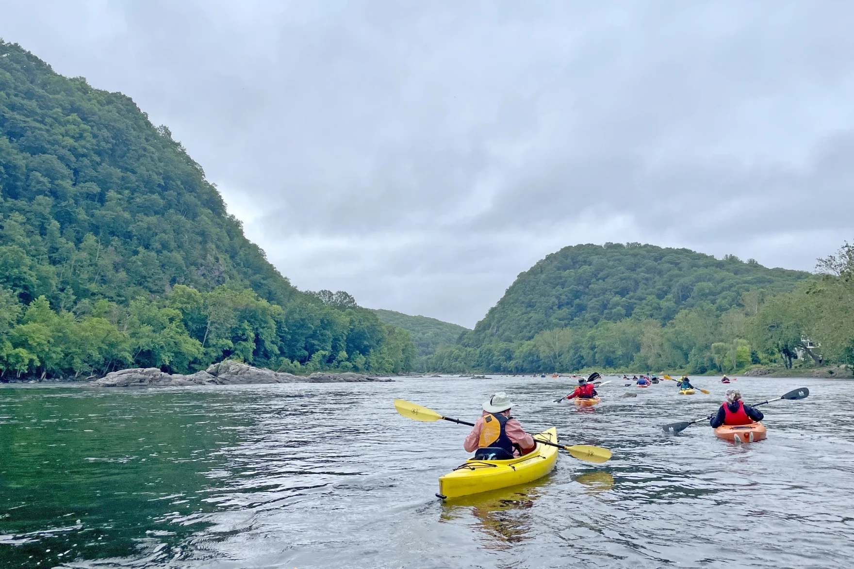 Kayakers paddle along a river flowing through wooded hillsides.