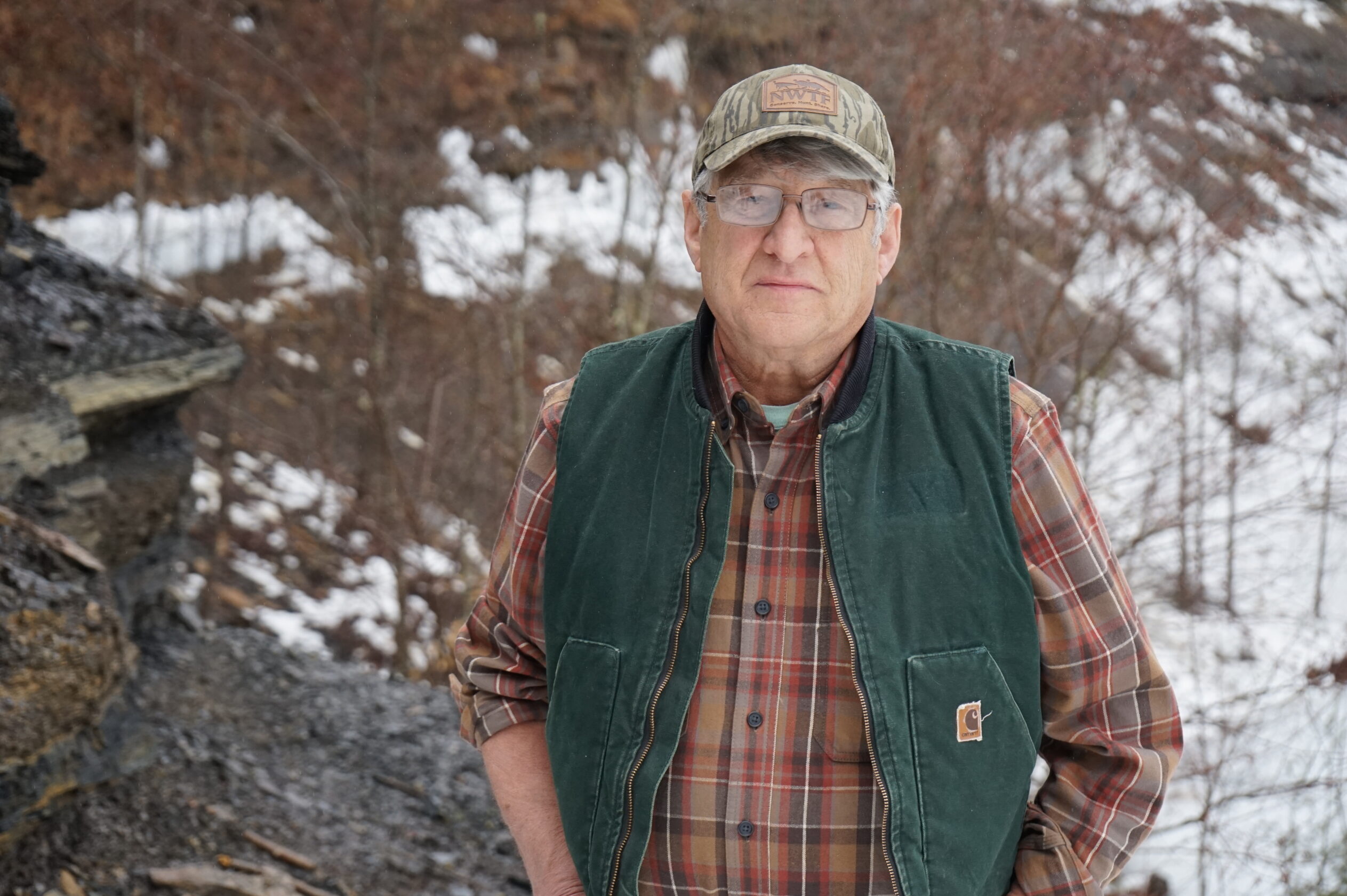 A man in a green vest and tan baseball hat stands in front of a snowy hill.