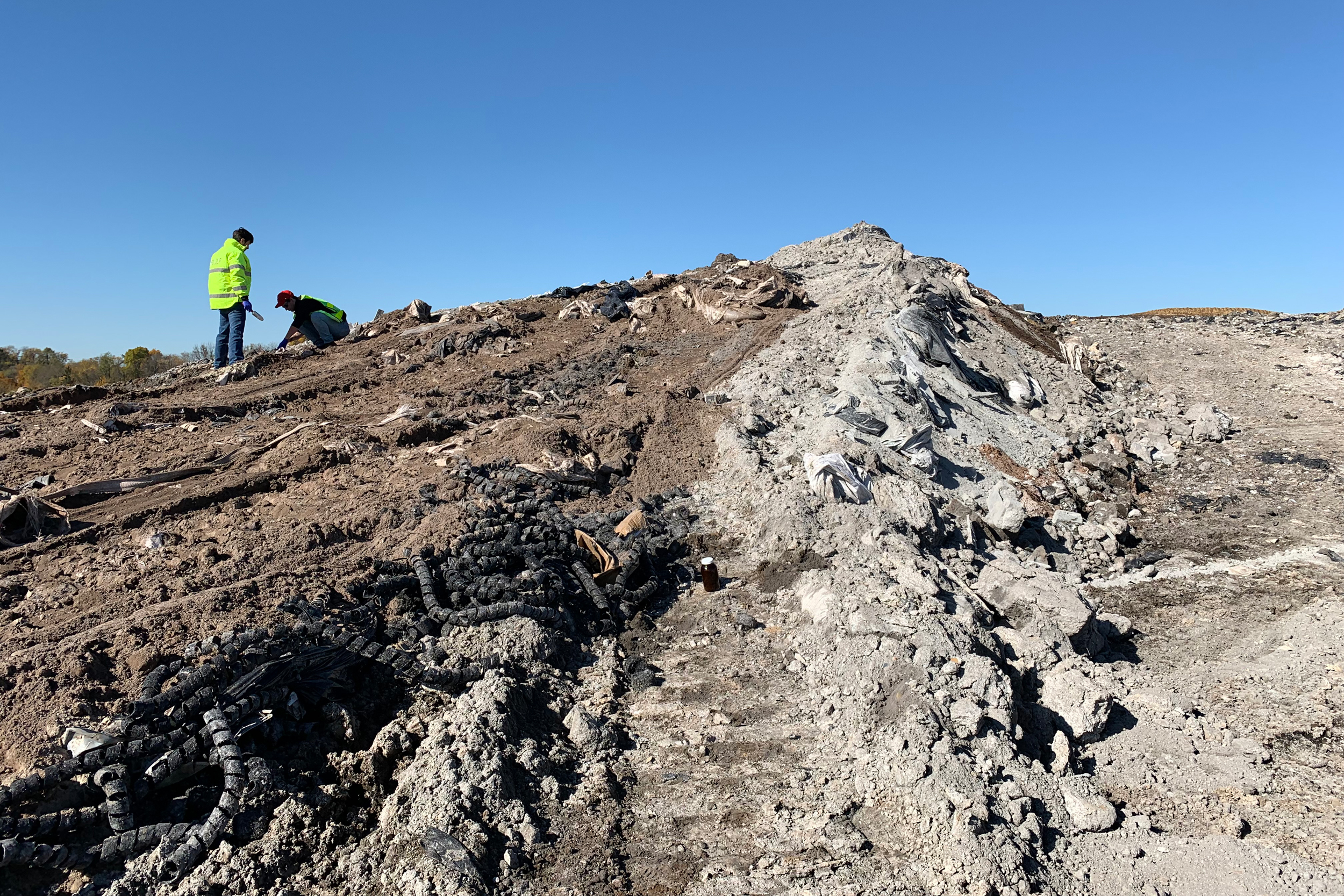 Two people in bright yellow safety coats stand at the top of a hill of exposed dirt and rocks with debris mixed in.