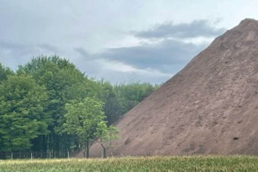 A huge pile of brown coal ash next to a wooded area and field. It is the size of a small hill,